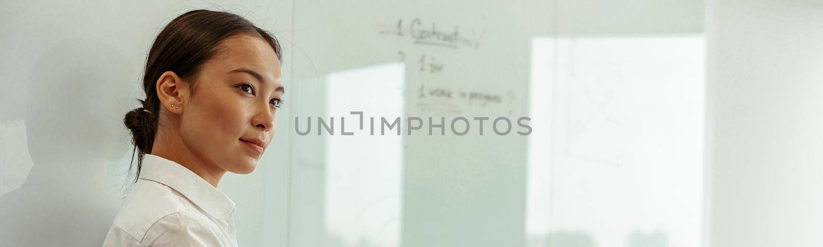 Portrait of asian business woman on background of project planning flipboard looks away by Yaroslav_astakhov