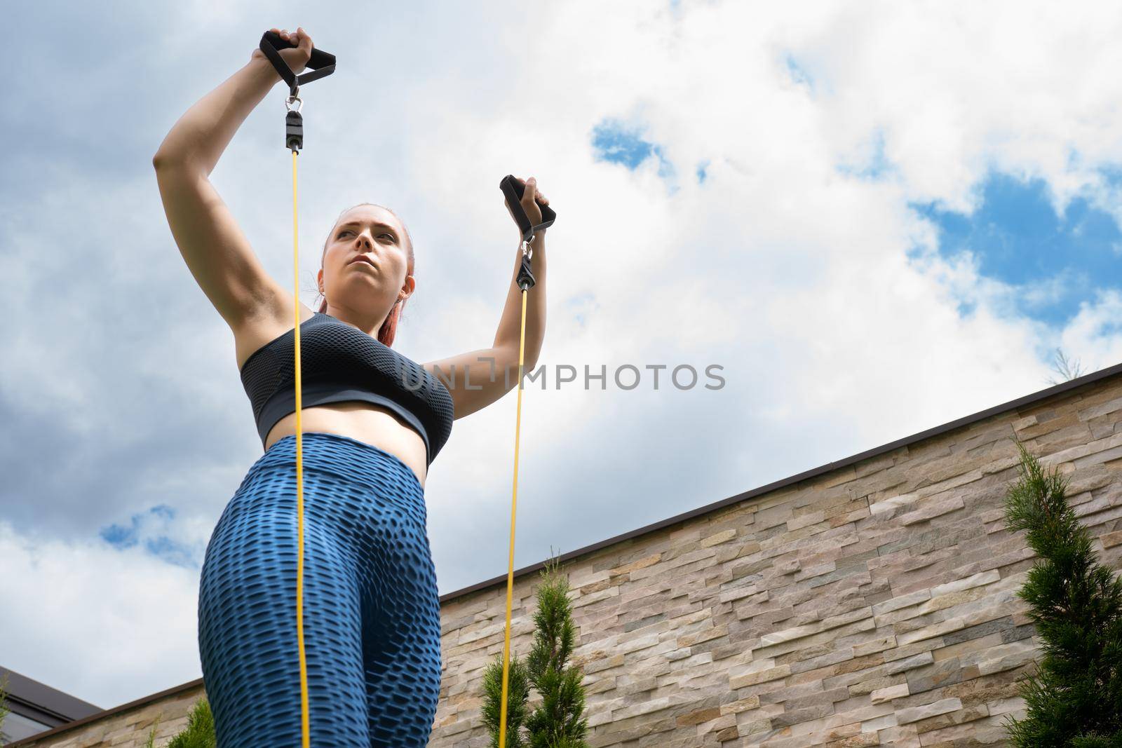 Attractive young fitness girl practising stretching exercise with rubber bands outdoors in the garden of her house. health and wellness concept.natural light in the garden.