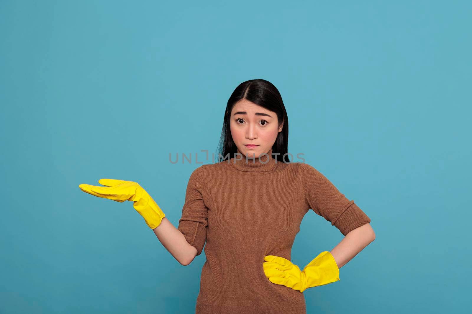 Asian woman with fearful facial expression pointing hand aside at copy space and wearing yellow gloves for safety, Housewife worker, Cleaning home concept, Stressed worried female