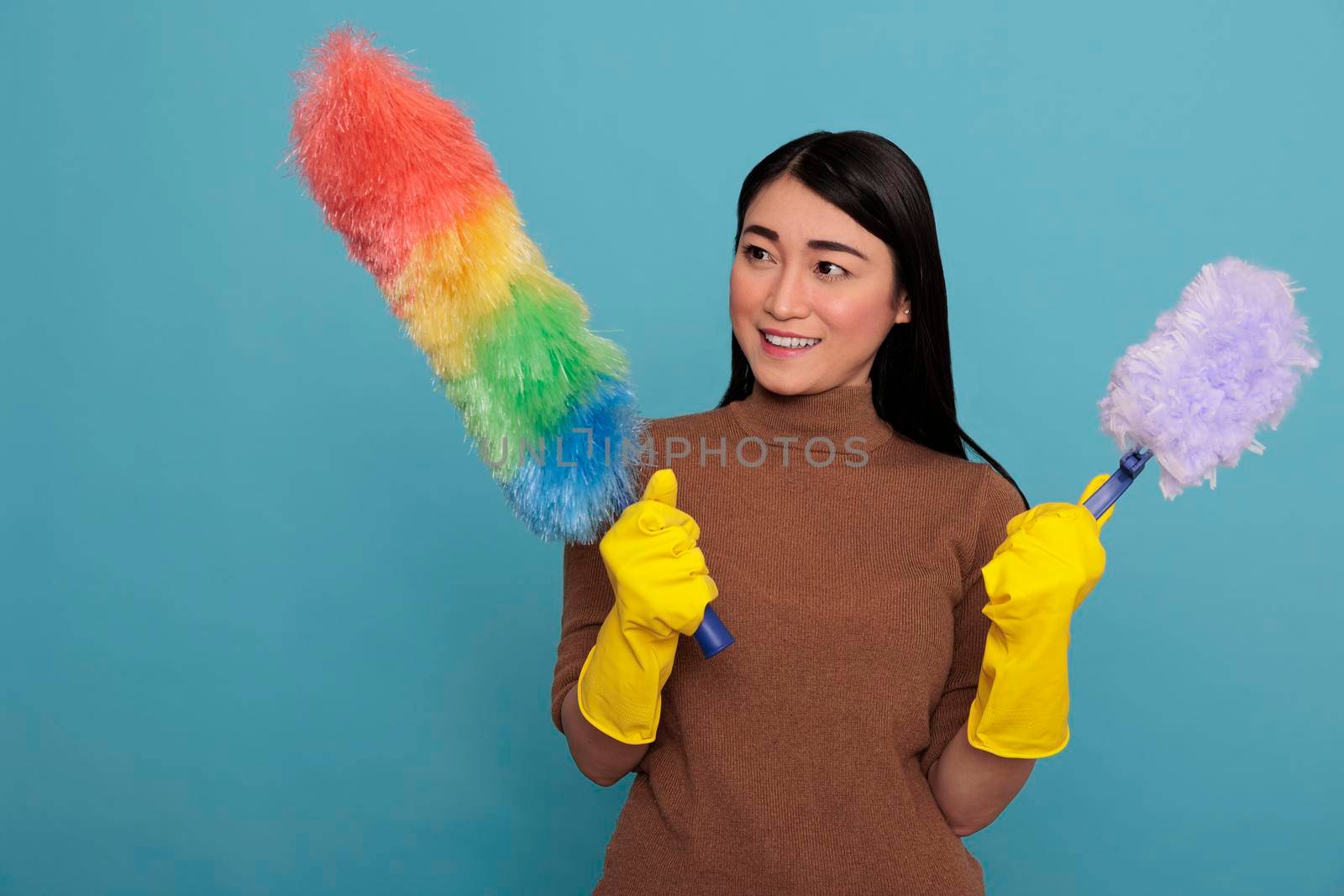 Cheerful happy and smiling asian young woman holding two duster brush in yellow gloves, Cleaning home concept, smiling female ready for his duty, Housewife feeling glad satisfied and positive