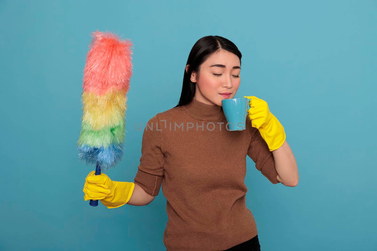 Portrait of young woman taking coffee for easy feel in the yellow glove with colorful duster brush on other hand isolated on a blue background, Cleaning home, Refreshment tea time