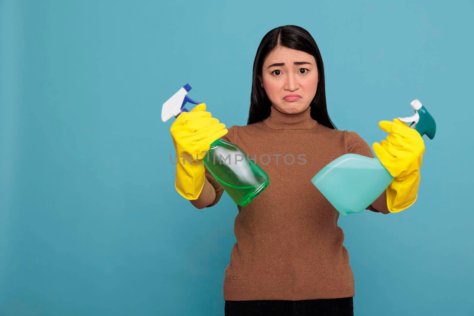 Sadly angry young asian woman holding two detergent spray in yellow gloves isolated on a blue background, Cleaning home concept, Unhappy depressed and exhausted negative housekeeper