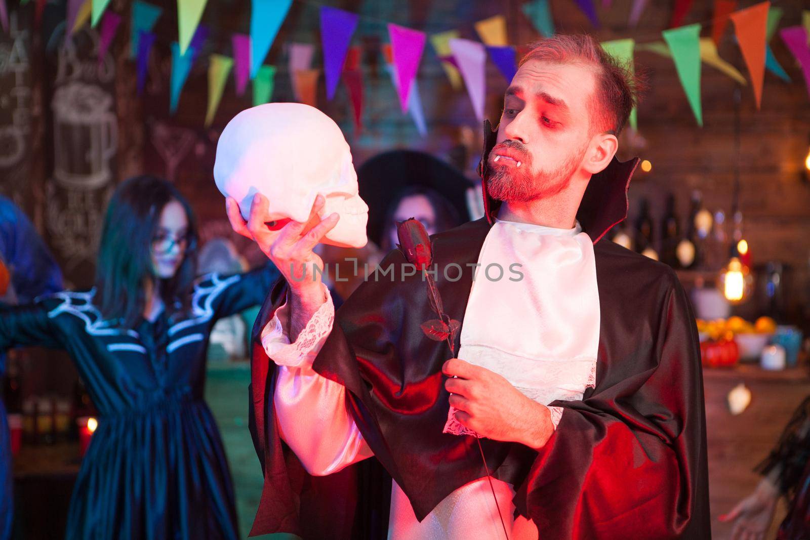Attractive young man dressed up like dracula holding a black rose looking at his human skull at halloween celebration by DCStudio