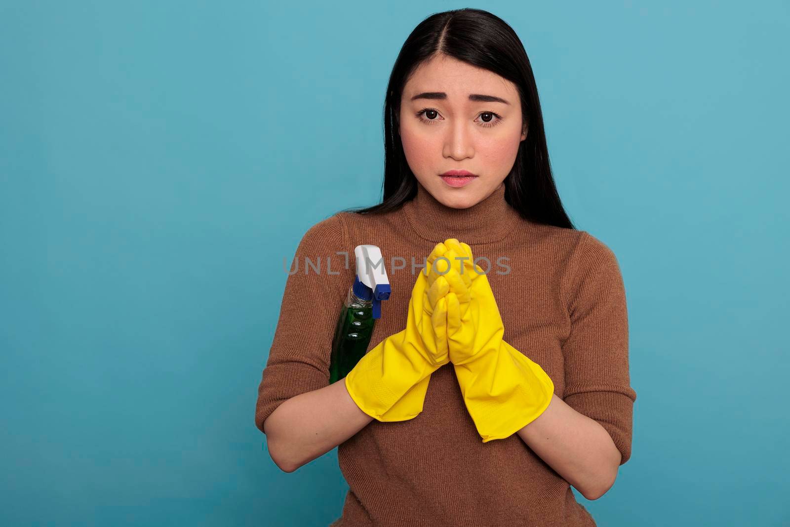 Young frightened scared asian housewife put her hands together wearing yellow gloves and a detergent sprayer under arm isolated against a blue background, Housewife worker, Cleaning home concept,