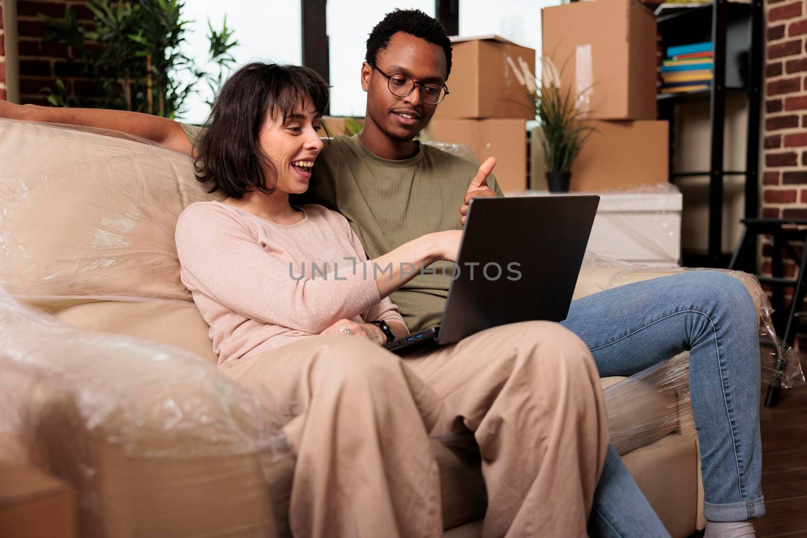 Diverse couple doing online shopping on laptop to buy furniture, browsing internet website for home decor inspiration. Moving in rented apartment property together for relationship event.