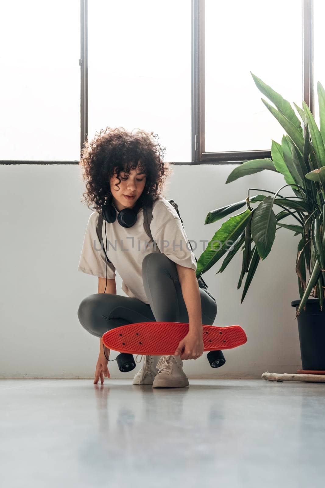 Young multiracial woman with curly hair crouching down holding a red skateboard looking down. by Hoverstock