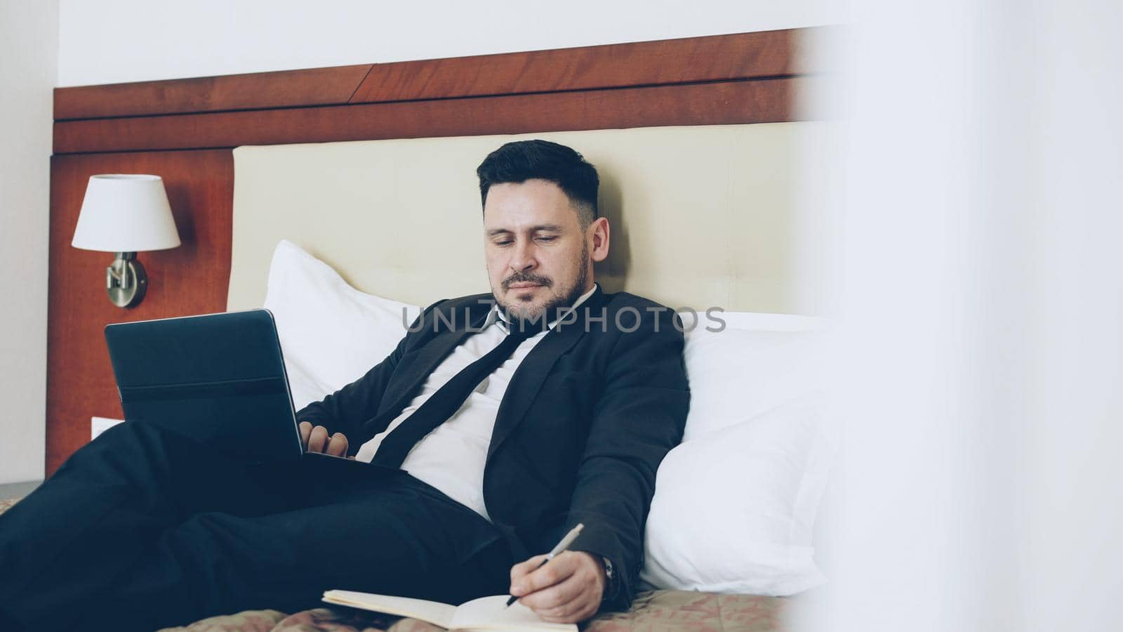 Confident bearded businessman lying on bed using laptop computer and writing notes at notepad while working in hotel room. Travel, business and people concept