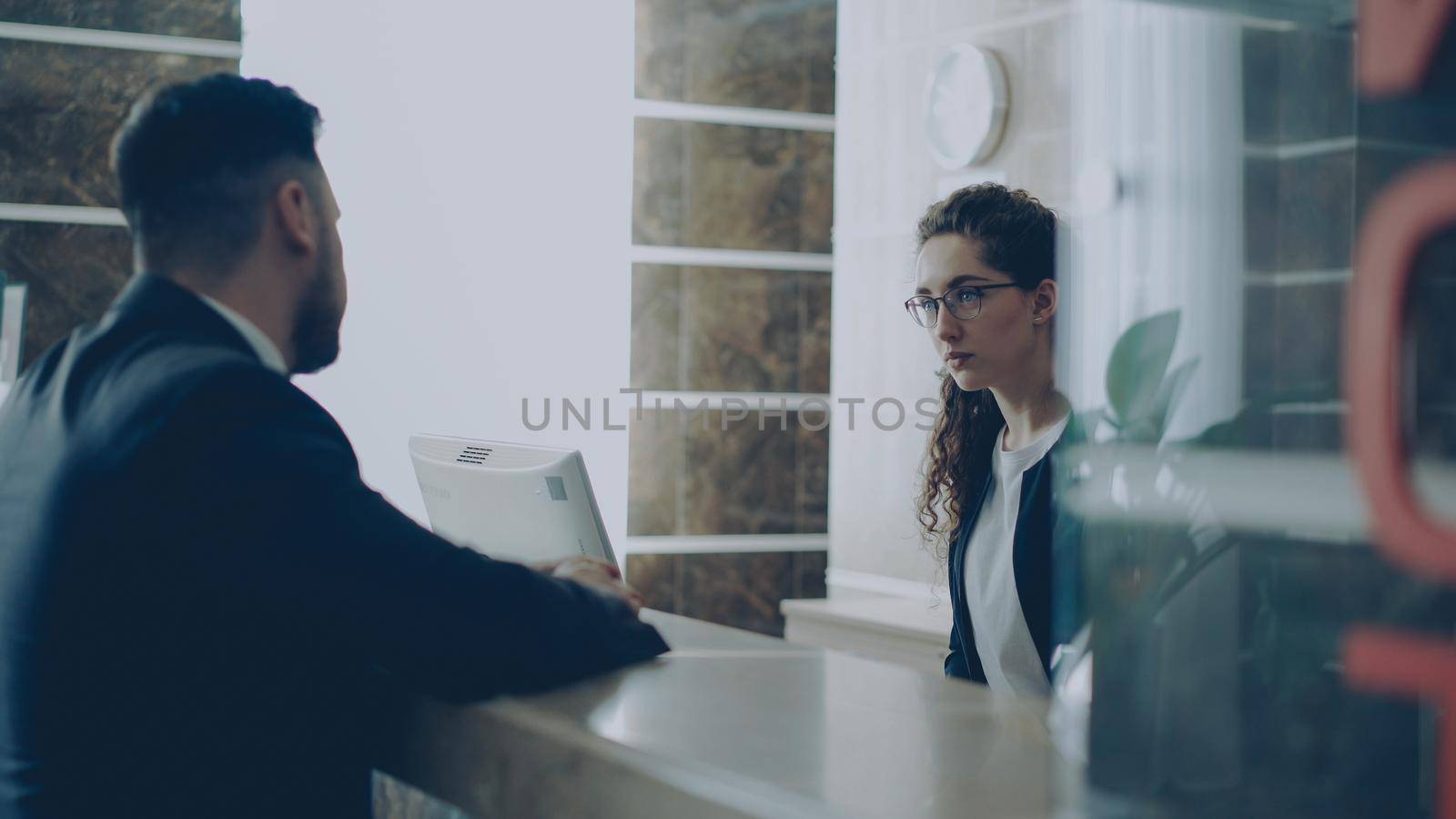 Receptionist girl talking with arrived businessman about check-in at reception in hotel. Business, travel and people concept by silverkblack