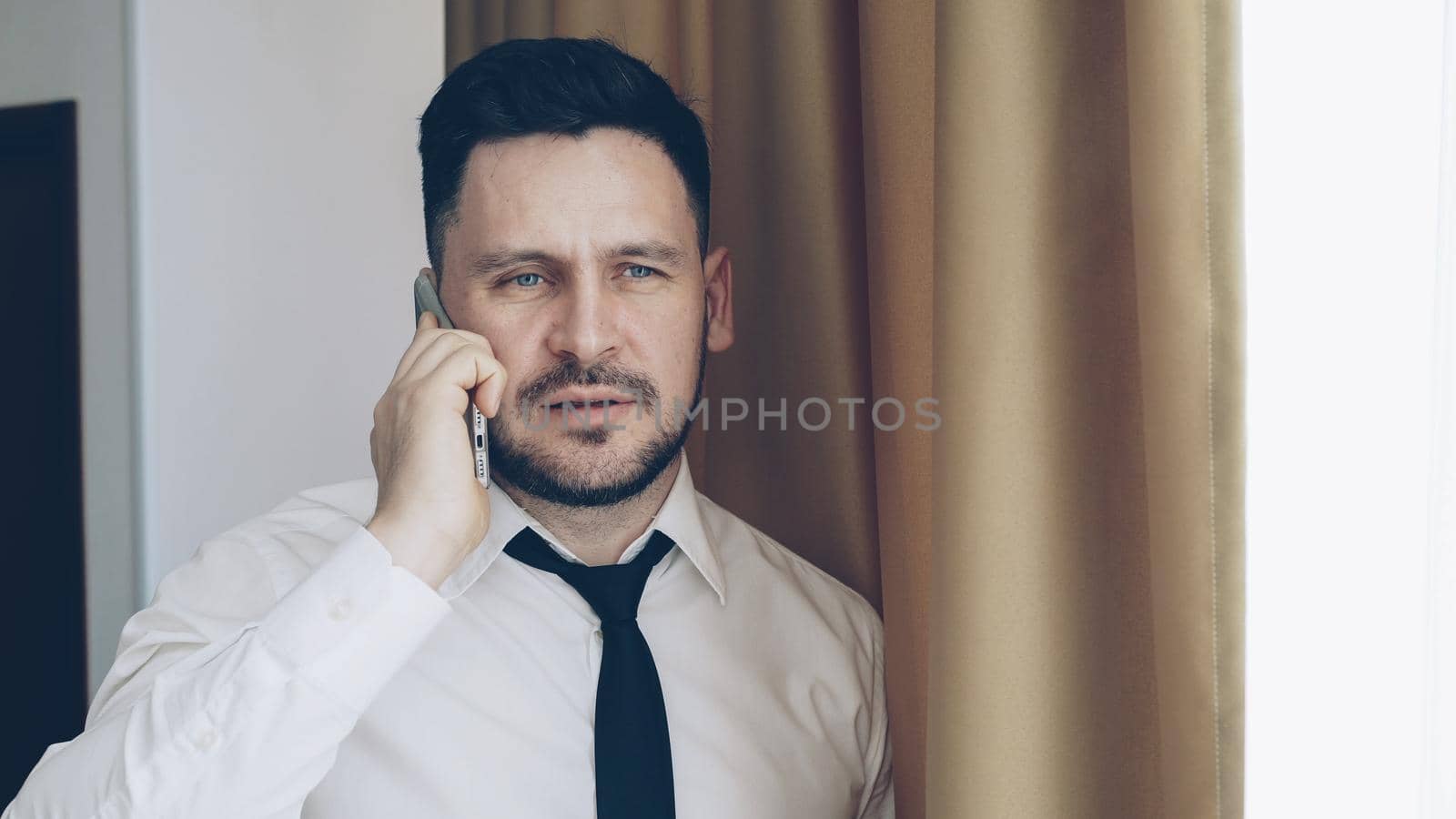 Adult bearded businessman in white shirt and tie talking on mobile phone standing near window in hotel room by silverkblack