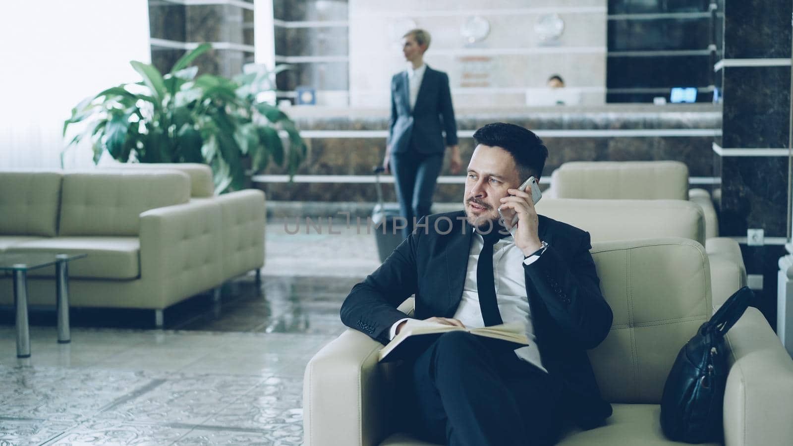 Pan shot of handsome bearded businessman sitting in armchair talking mobile phone with notepad while businesswoman in suit with luggage walking through hotel lobby from reception desk.