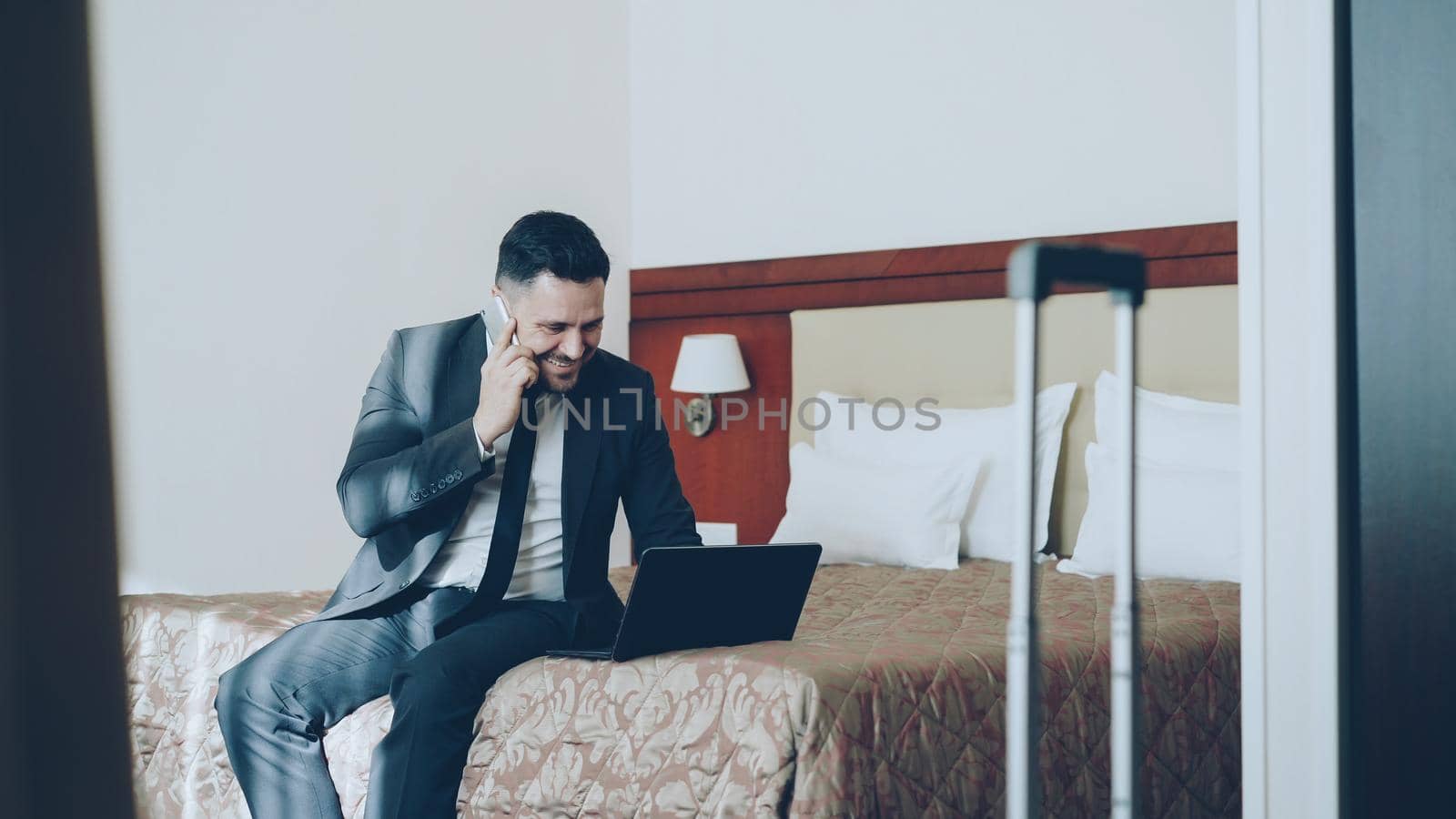 Tilt up of busy businessman in suit working on laptop and talking at mobile phone while sitting on bed in hotel room. Business, travel and people concept by silverkblack