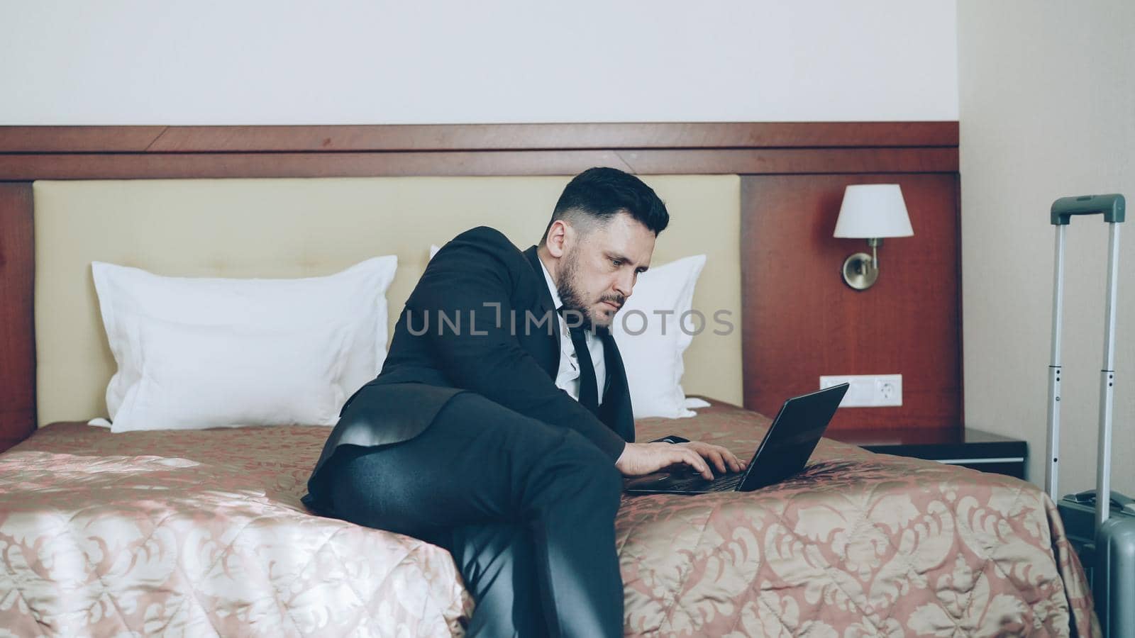 Pan shot of concentrated young businessman in suit working on laptop while sitting on bed in hotel room. Business, travel and people concept