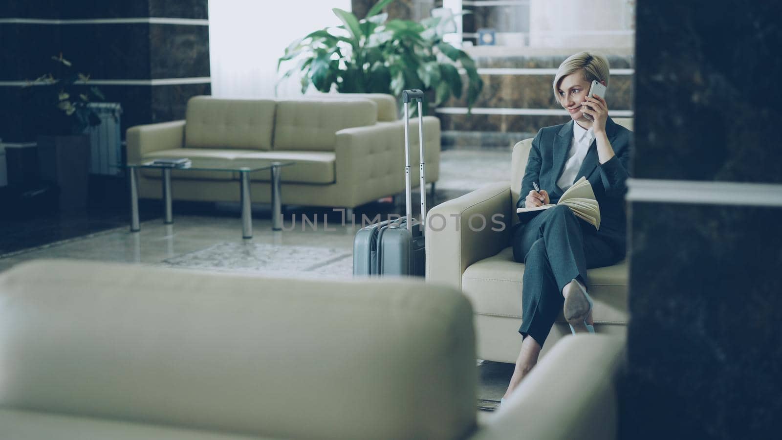 Cheerful blonde businesswoman sitting in armchair in hotel lobby talking on mobile phone and writing in notepad smiling and looking aside. Business, travel and people concept by silverkblack