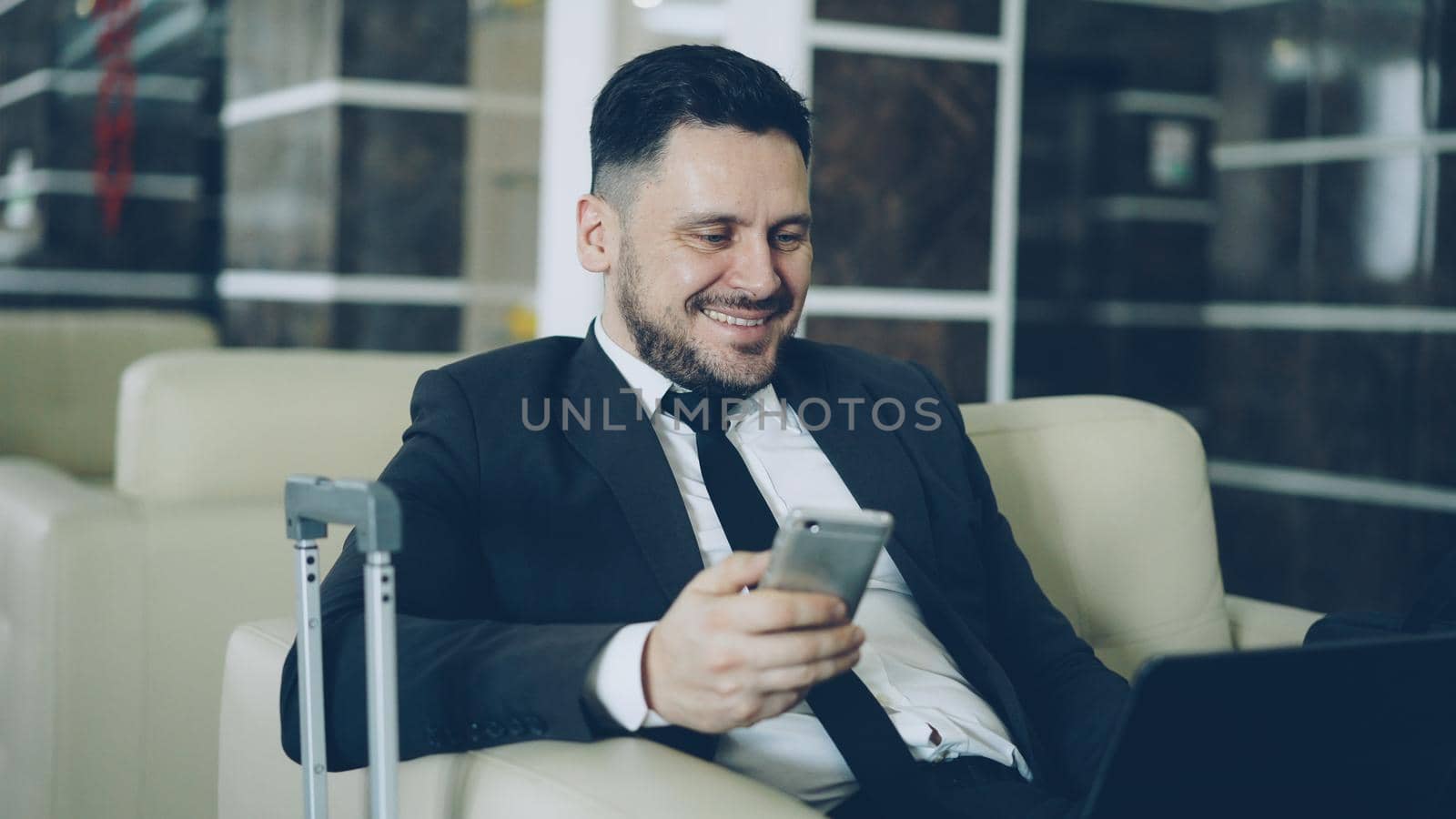 Handsome young businessman using smarphone and laptop computer while working in comfortable hotel lobby with luggage suitcase near him. Travel, business and people concept