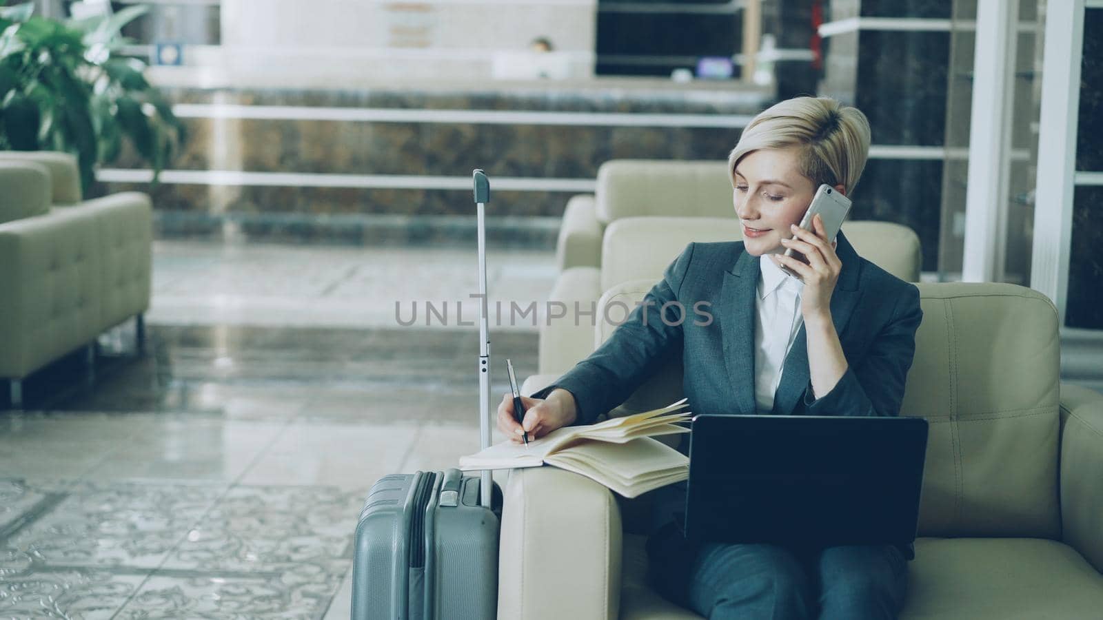 Pan shot of blonde busy businesswoman sitting in armchair in hotel lobby talking mobile phone, writing in notepad and using laptop computer. Business, travel and people concept by silverkblack