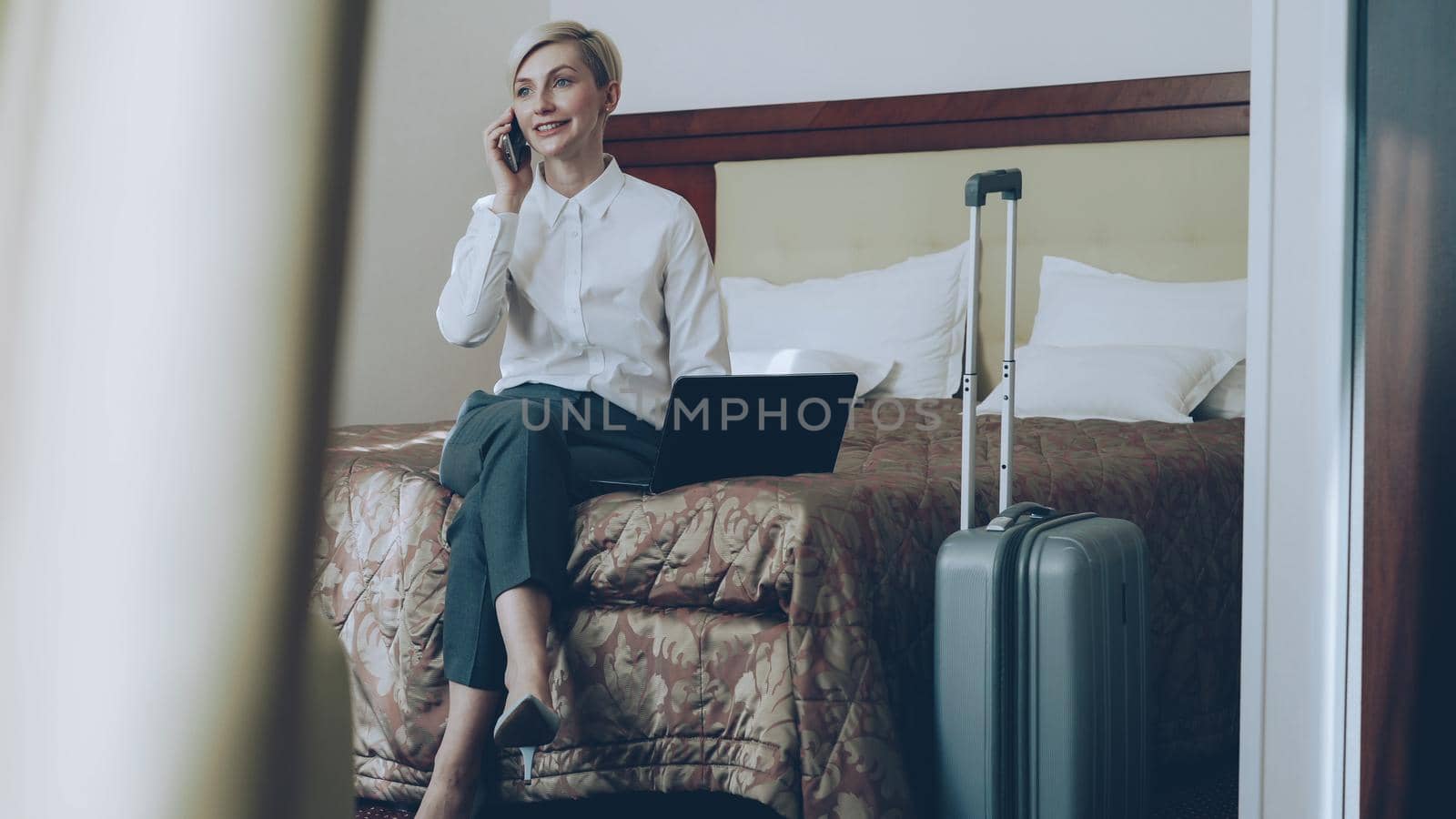 PAN of smiling businesswoman in white shirt using on laptop and talking at mobile phone while sitting on bed in hotel room. Business, travel and people concept by silverkblack