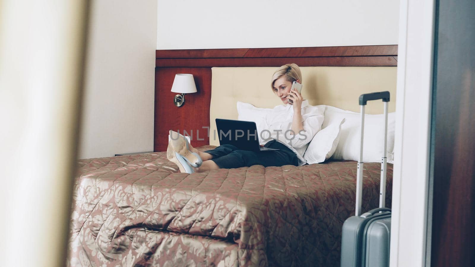 Smiling businesswoman in white shirt using on laptop and talking at mobile phone while lying on bed in hotel room. Business, travel and people concept by silverkblack