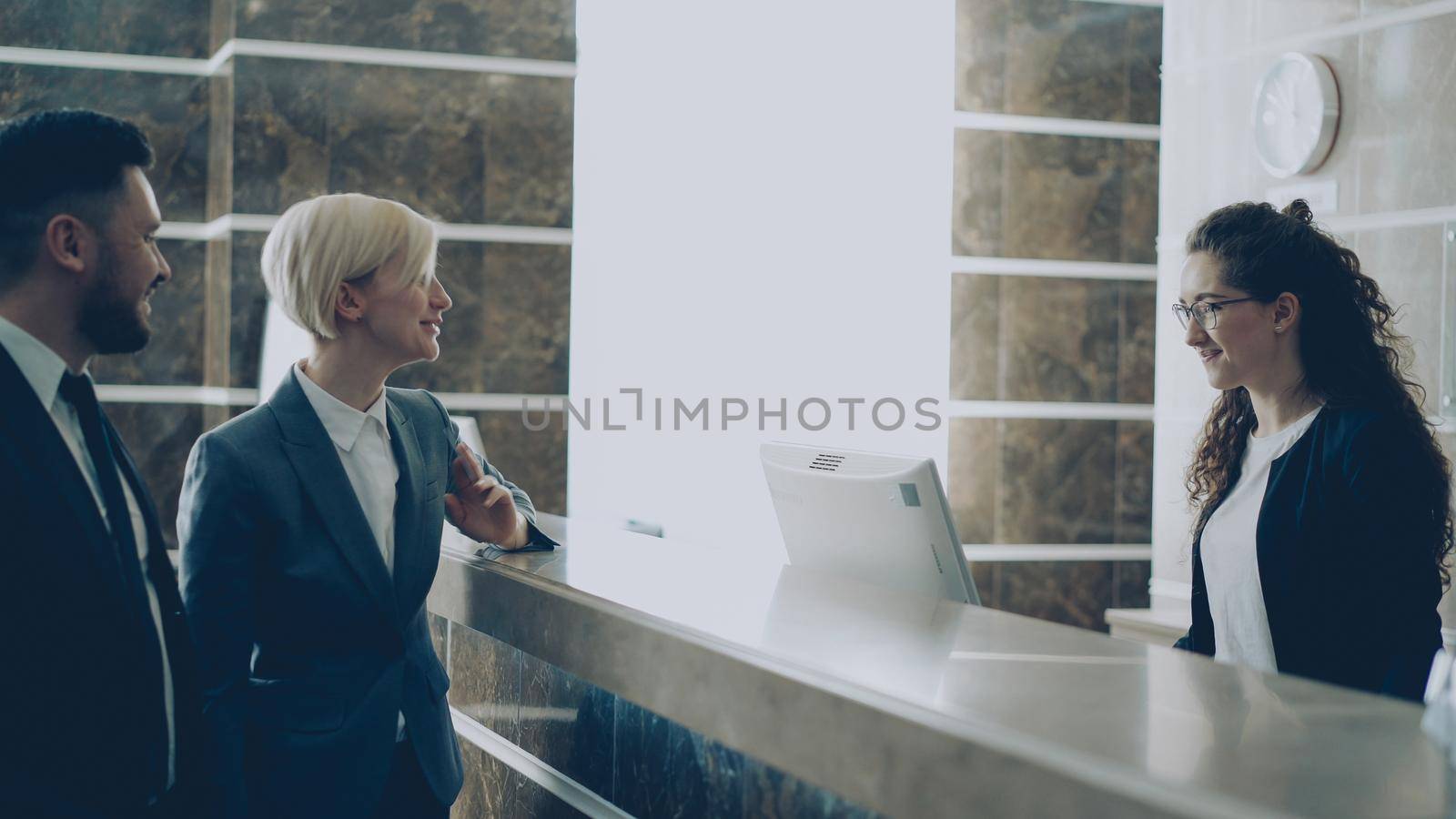 Receptionist girl talking with arrived businessman and businesswoman guests about check-in in hotel and giving key card from room to man . Business, travel and people concept by silverkblack