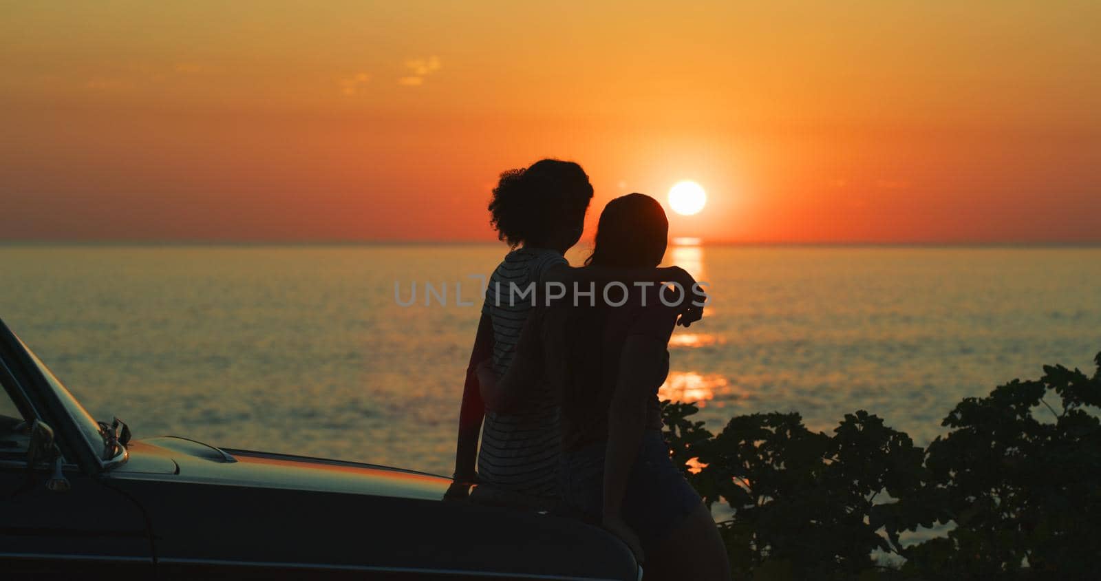Watching the sun slowly kiss the sea. two unrecognizable girl friends watching the sunset while on a road trip at the beach