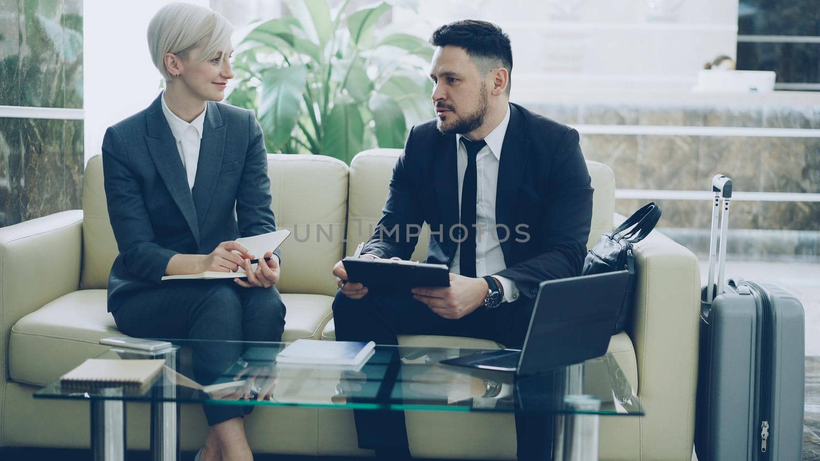 Two female and male business colleagues with clipboard and notepad talking at hotel lobby. Luggage in near couch. Business, travel and people concept by silverkblack