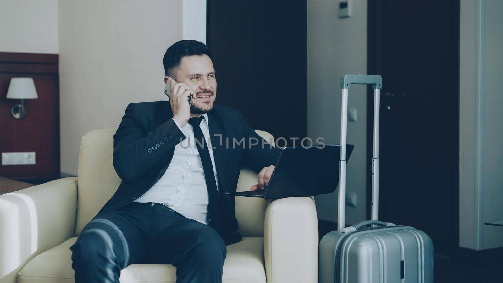 Confident cheerful businessman in suit checking laptop computer and talking at mobile phone while sitting in armchair at hotel room. Business, travel and people concept