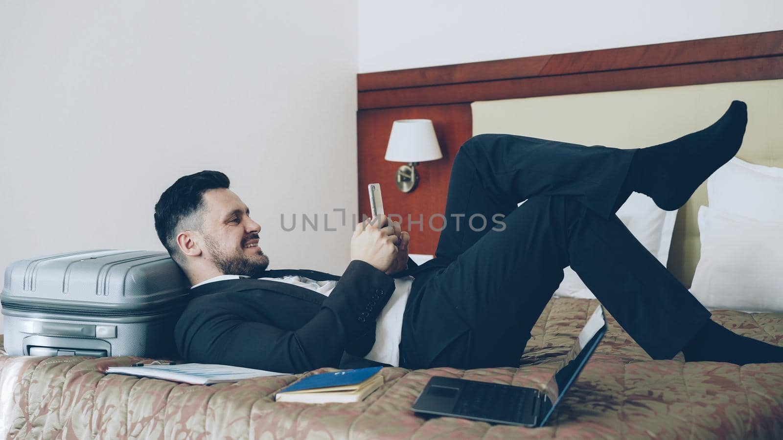 Young businessman lying on bed and suitcase using smartphone while resting after arrival at hotel room. Travel, business and people concept by silverkblack