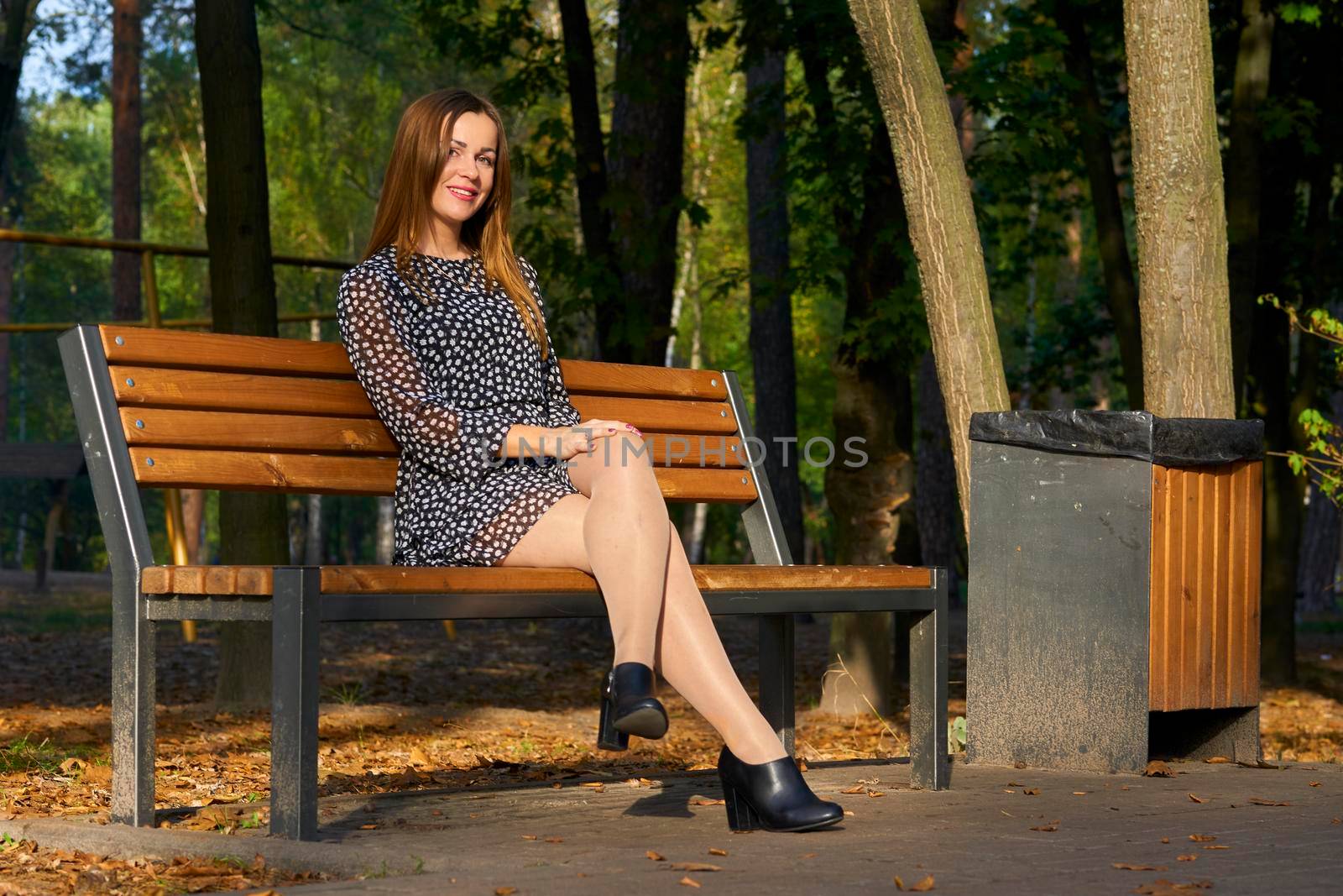 Young cute woman in a dress is resting in the autumn city park on a wooden bench by jovani68