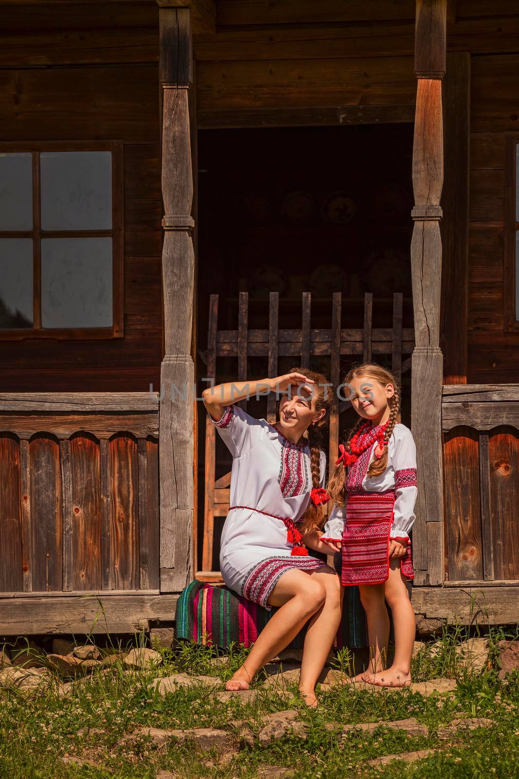 mother and daughter in Ukrainian national costumes are sitting near an old house