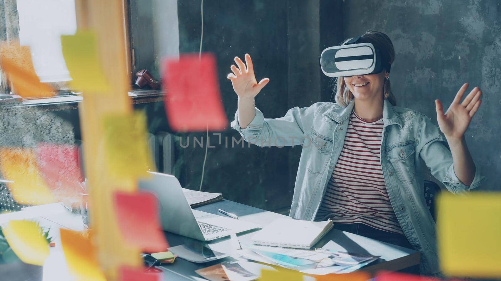 Attractive young woman is sitting at table and wearing virtual reality glasses. She is moving hands and and having VR experience in modern lof office indoors