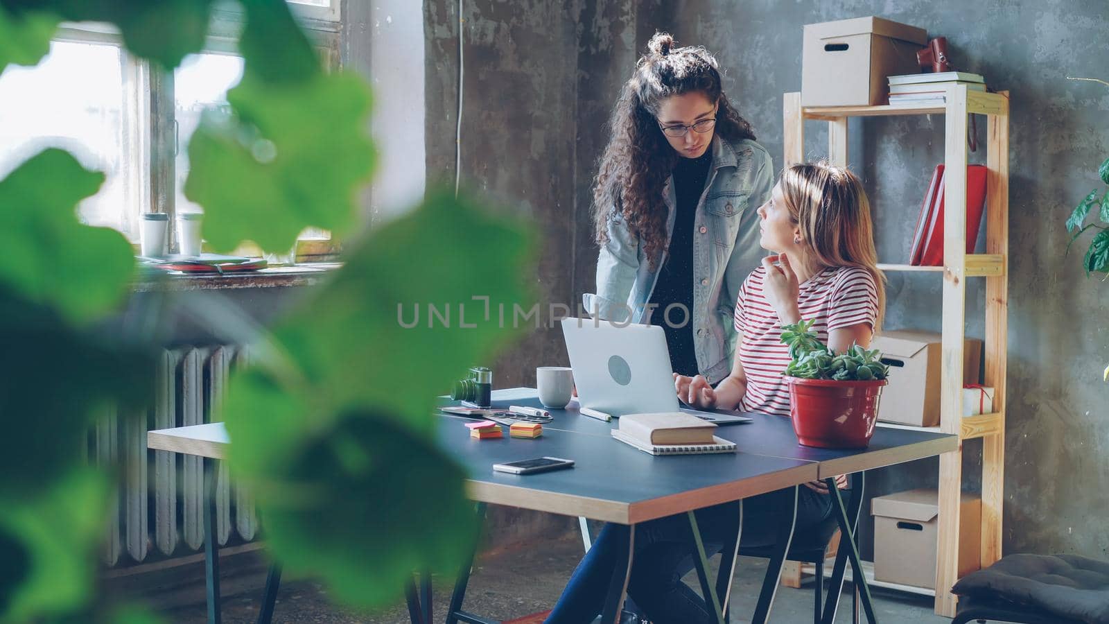 Young owners of small business are working with laptop in modern loft style office. Blonde is sitting and typing, brunette is standing near the desk and suggesting ideas looking at screen.
