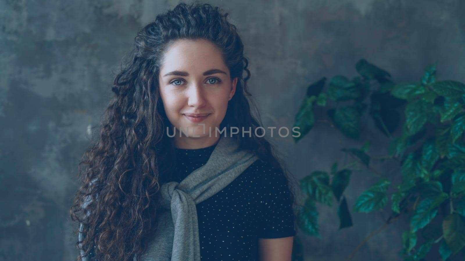 Close-up portrait of yougn attractive girl looking into camera and smiling while standing against grey wall and large green plant by silverkblack