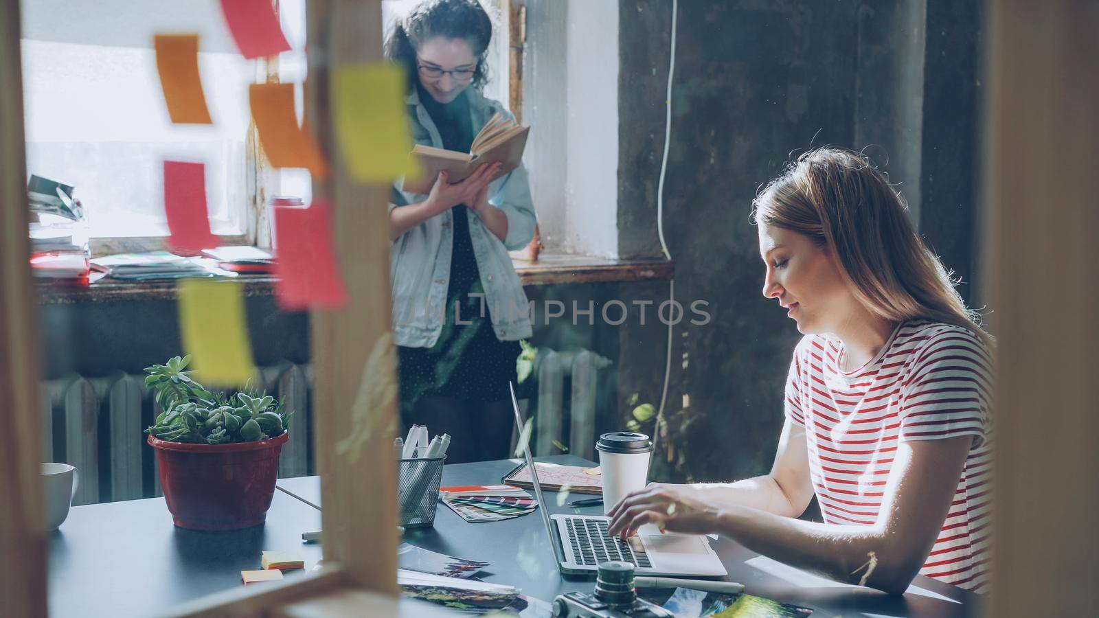 Young blond student wearing casual clothes is busy typing on laptop while her friend is reading book standing near large window. Loft style apartment is in background. Smart youth concept.