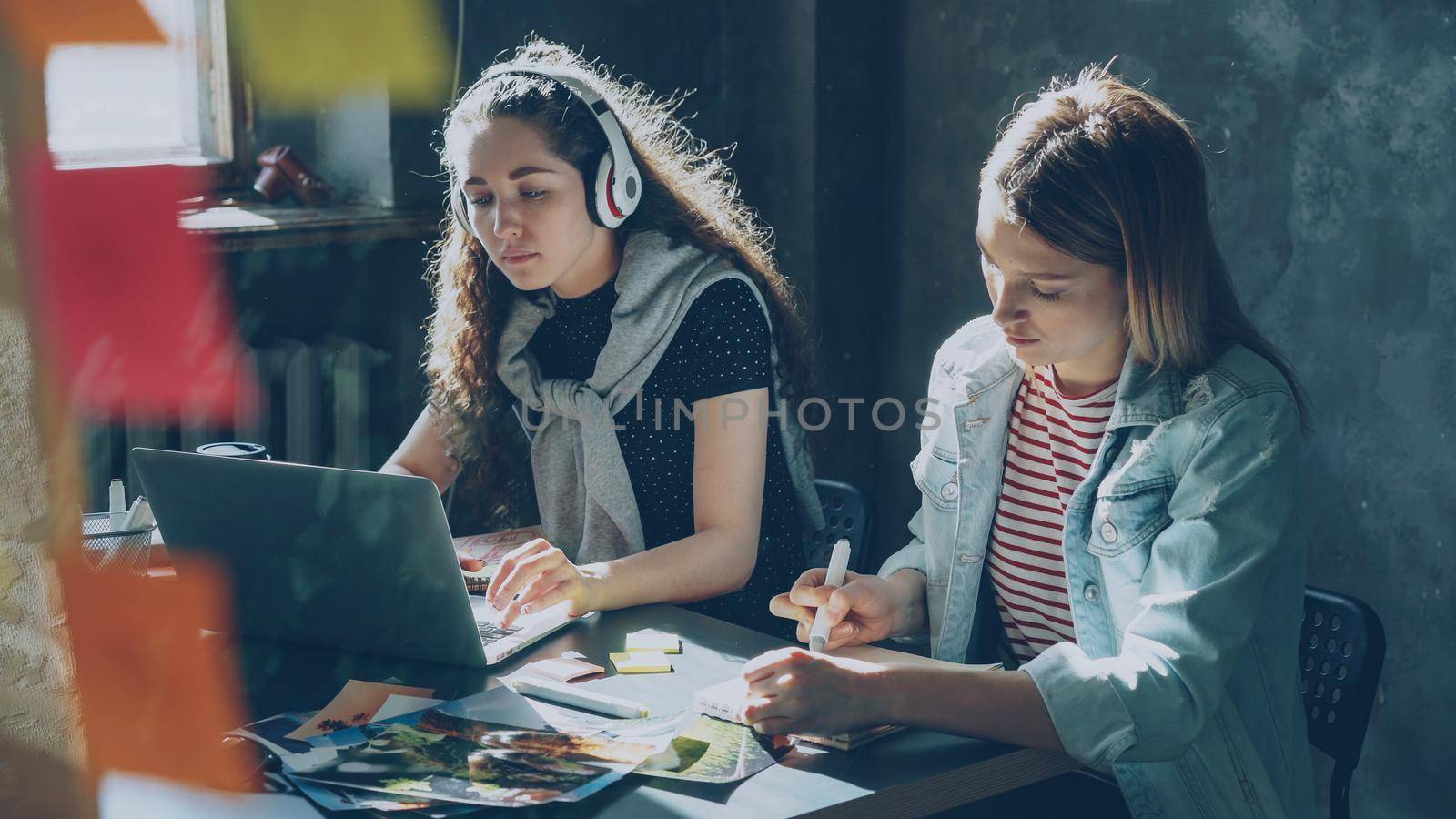 Team of creative designers is working together in light office. Cheerful dark-haired lady is listening to music and working with laptop, and blond woman is drawing images in notepad