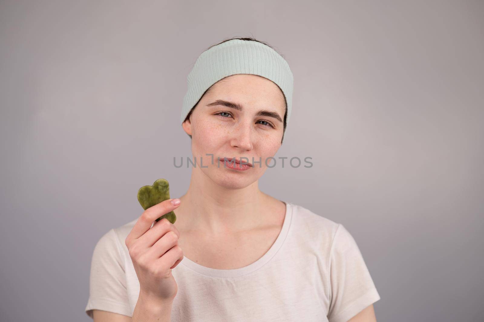 Portrait of a young woman massages her face with a gouache scraper on a white background