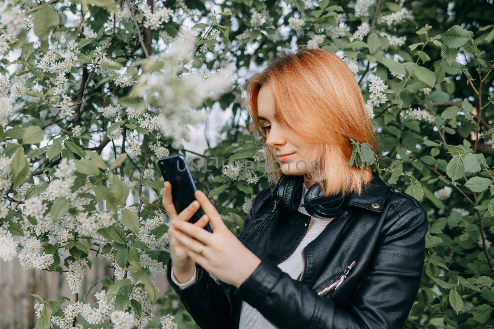 A red-haired teenage girl with headphones around her neck takes a selfie against the background of a flowering cherry tree. The time of spring flowering of cherry orchards.