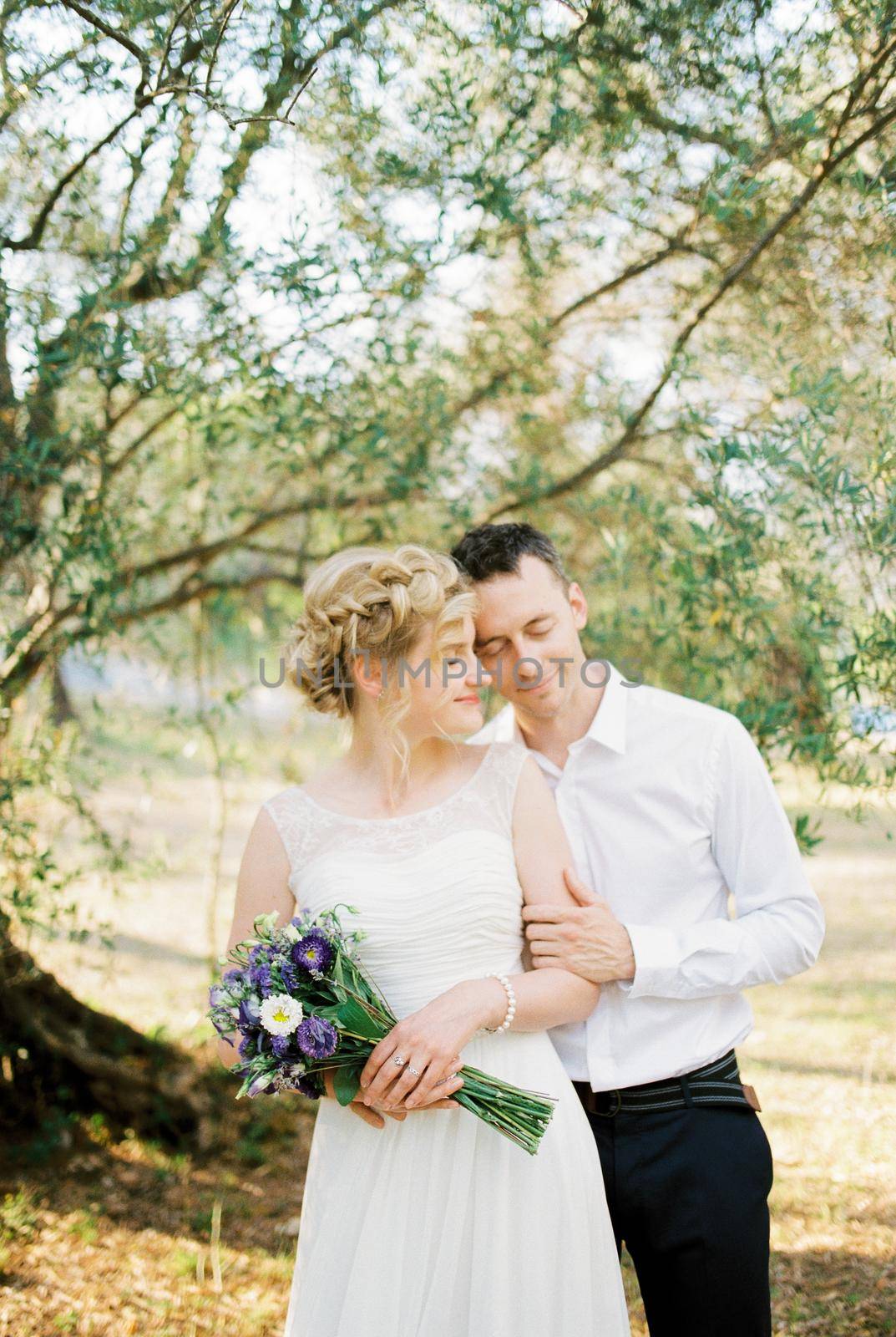 Groom hugs bride from behind with a bouquet near the olive tree. High quality photo