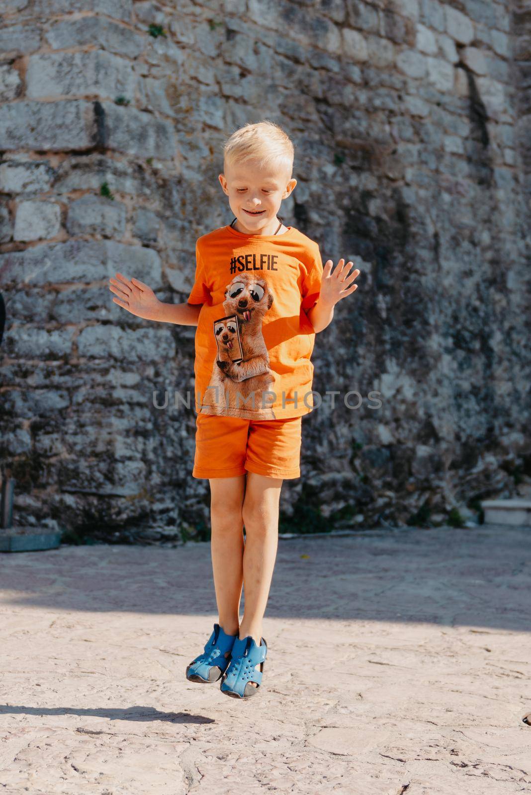 Kid's outdoor activity. Smile toddler boy wearing a orange shorts Jumping, running and having fun in a backyard on a sunny hot summer day. Full length of energetic little boy in stylish casual outfit jumping outdoor