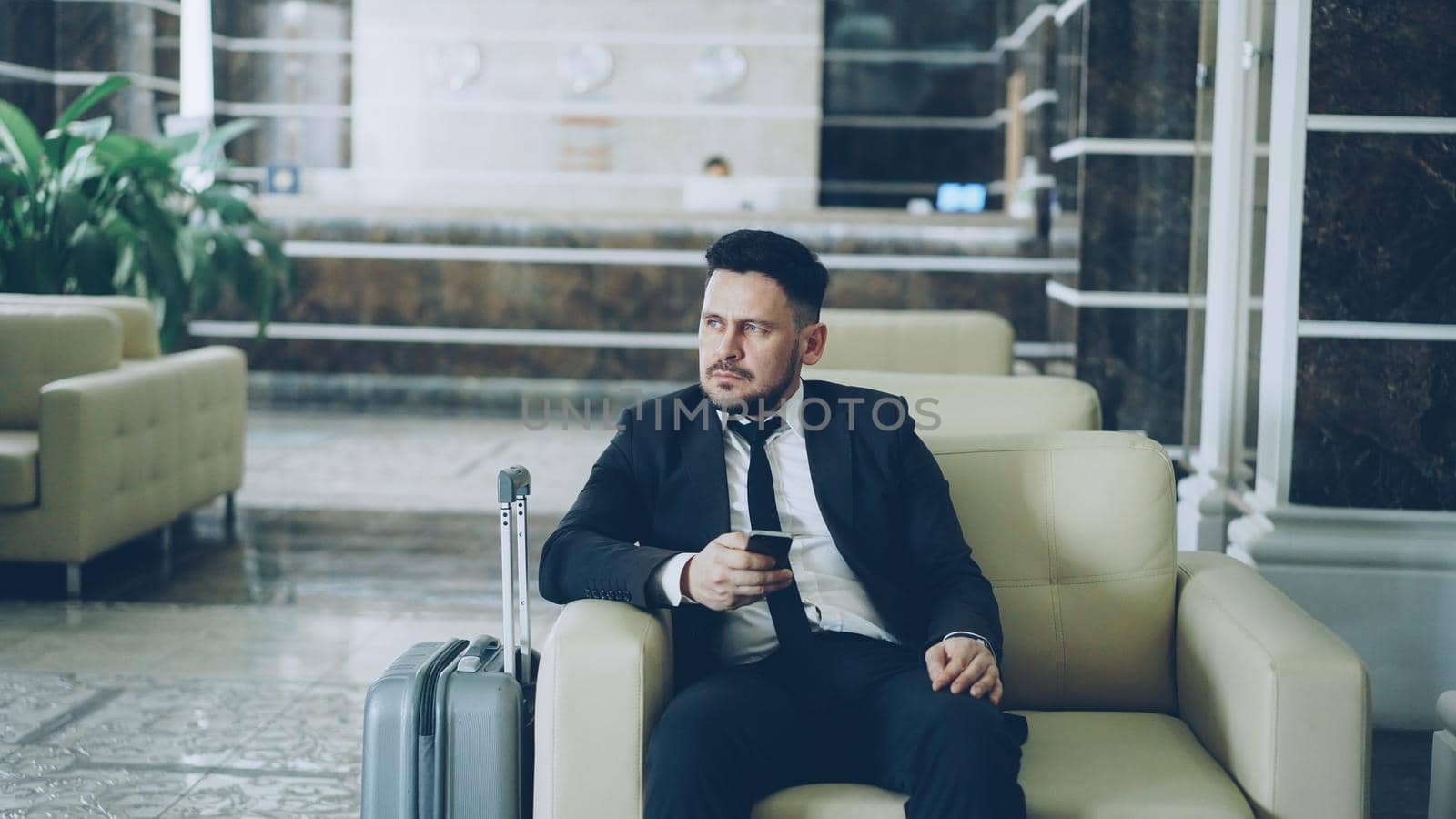 Pan shot of thoughtful businessman looking aside and using smartphone sitting on armchair in luxury hotel with luggage near him. Travel, business and people concept by silverkblack