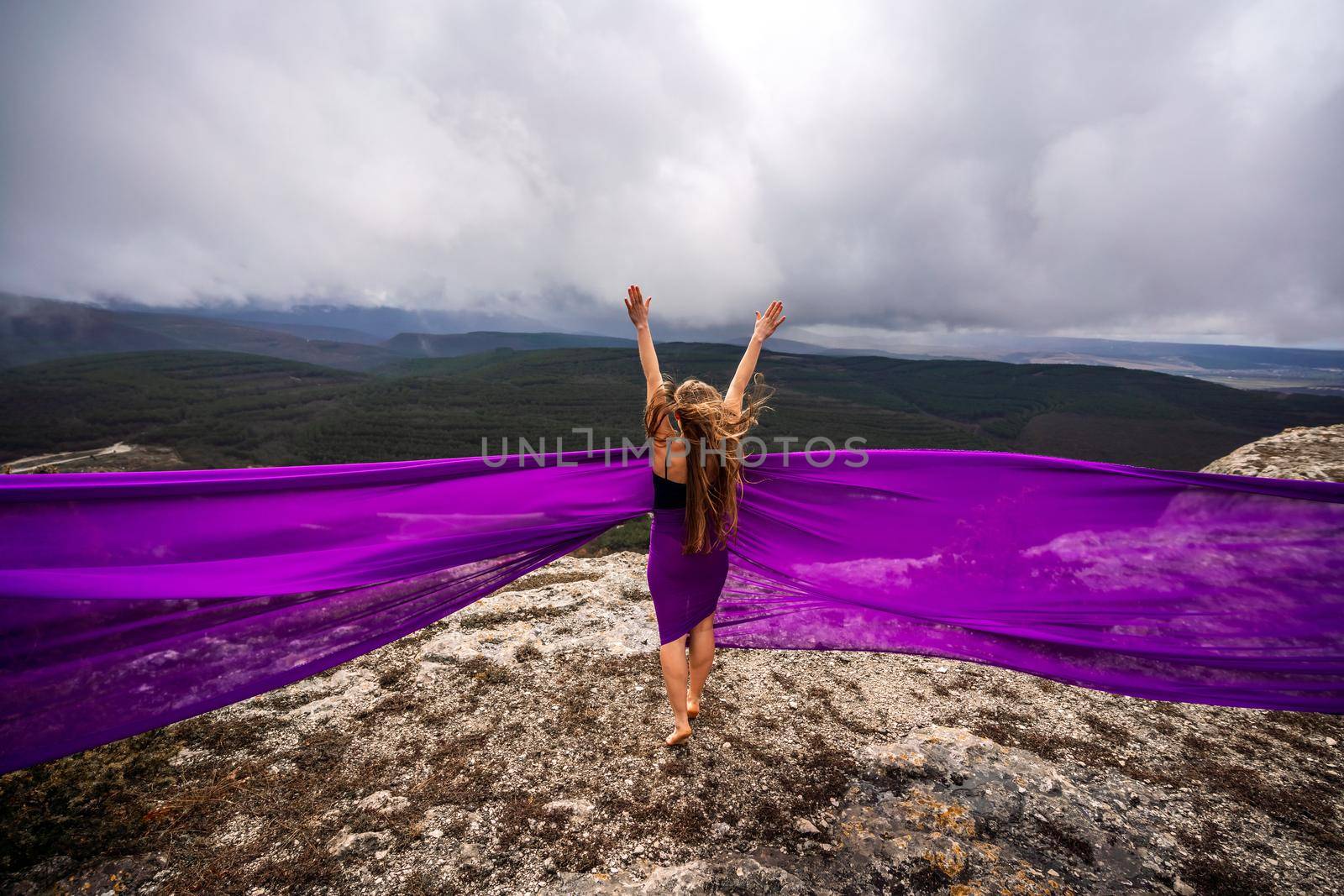 A woman with long hair is standing in a purple flowing dress with a flowing fabric. On the mountain against the background of the sky with clouds