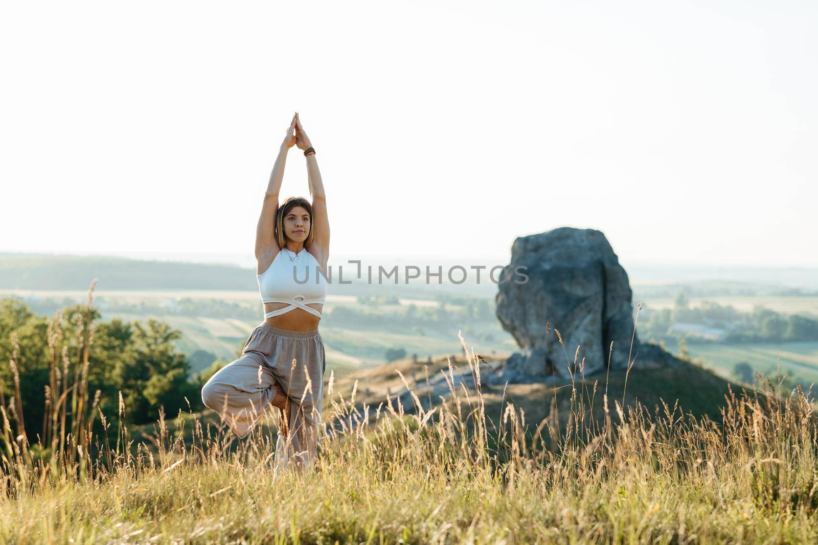 Young Woman Meditating and Practicing Yoga Outdoors at Sunset with Scenic Landscape and Nature Miracle Giant Stone on the Background by Romvy