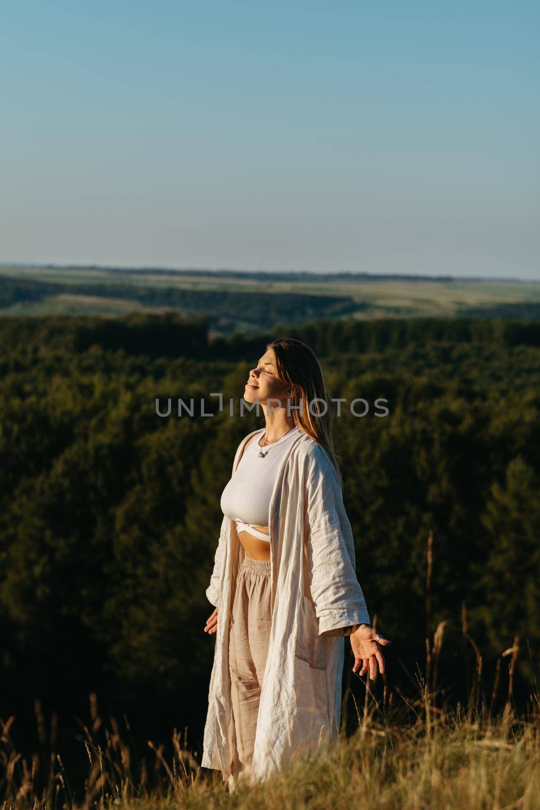 Young Woman Meditating and Catching Sunlight Outdoors at Sunset with Scenic Landscape on Background