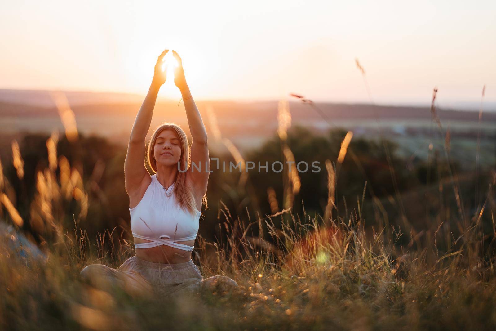 Young Woman Sitting in Meditation Yoga Pose and Catching Sun by Hands at Sunset by Romvy