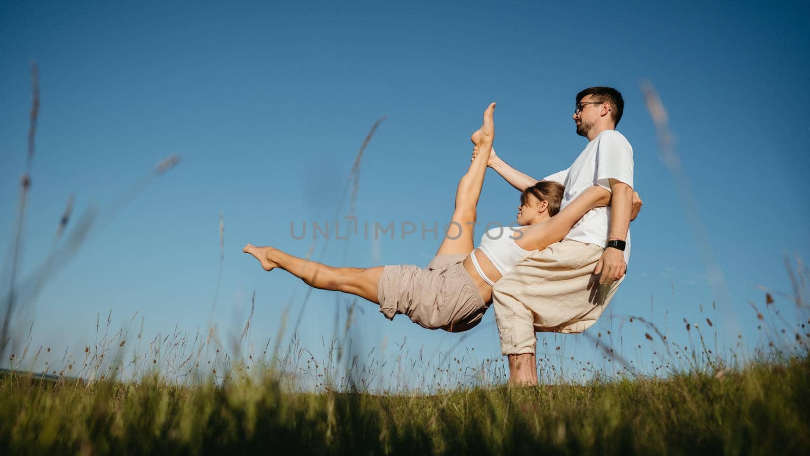 Man and Woman Dressed Alike Doing Difficult Pose While Practicing Yoga Outdoors in the Field with Blue Sky on the Background by Romvy