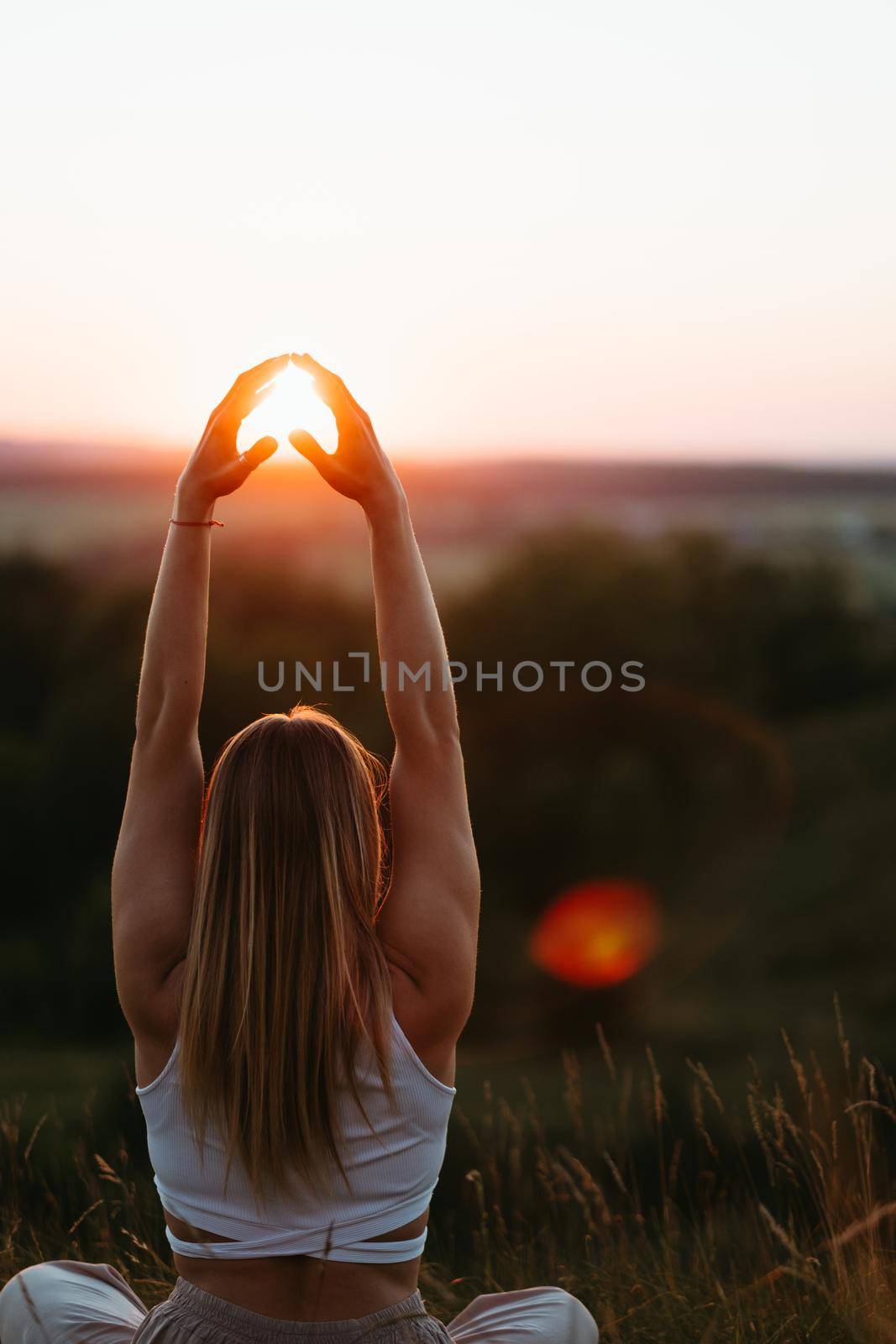 Back View on Woman Sitting in Meditation Yoga Pose and Catching Sun by Her Hands at Sunset Outdoors