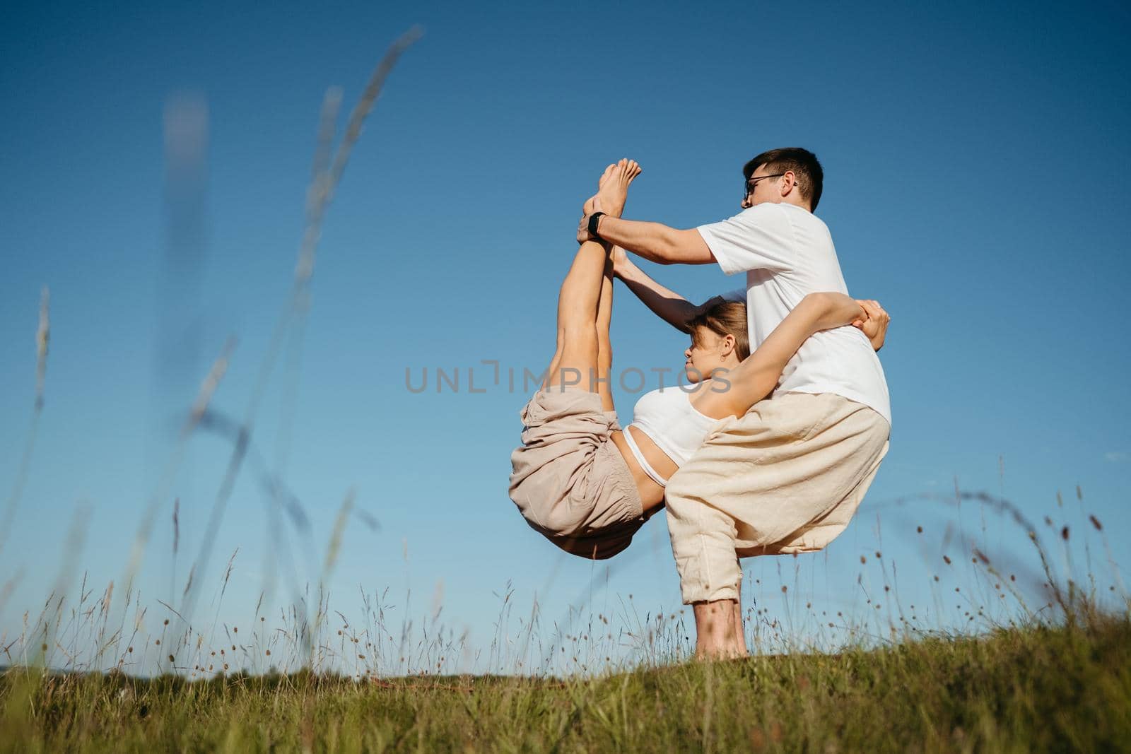 Man and Woman Dressed Alike Doing Difficult Pose While Practicing Yoga Outdoors in the Field with Blue Sky on Background