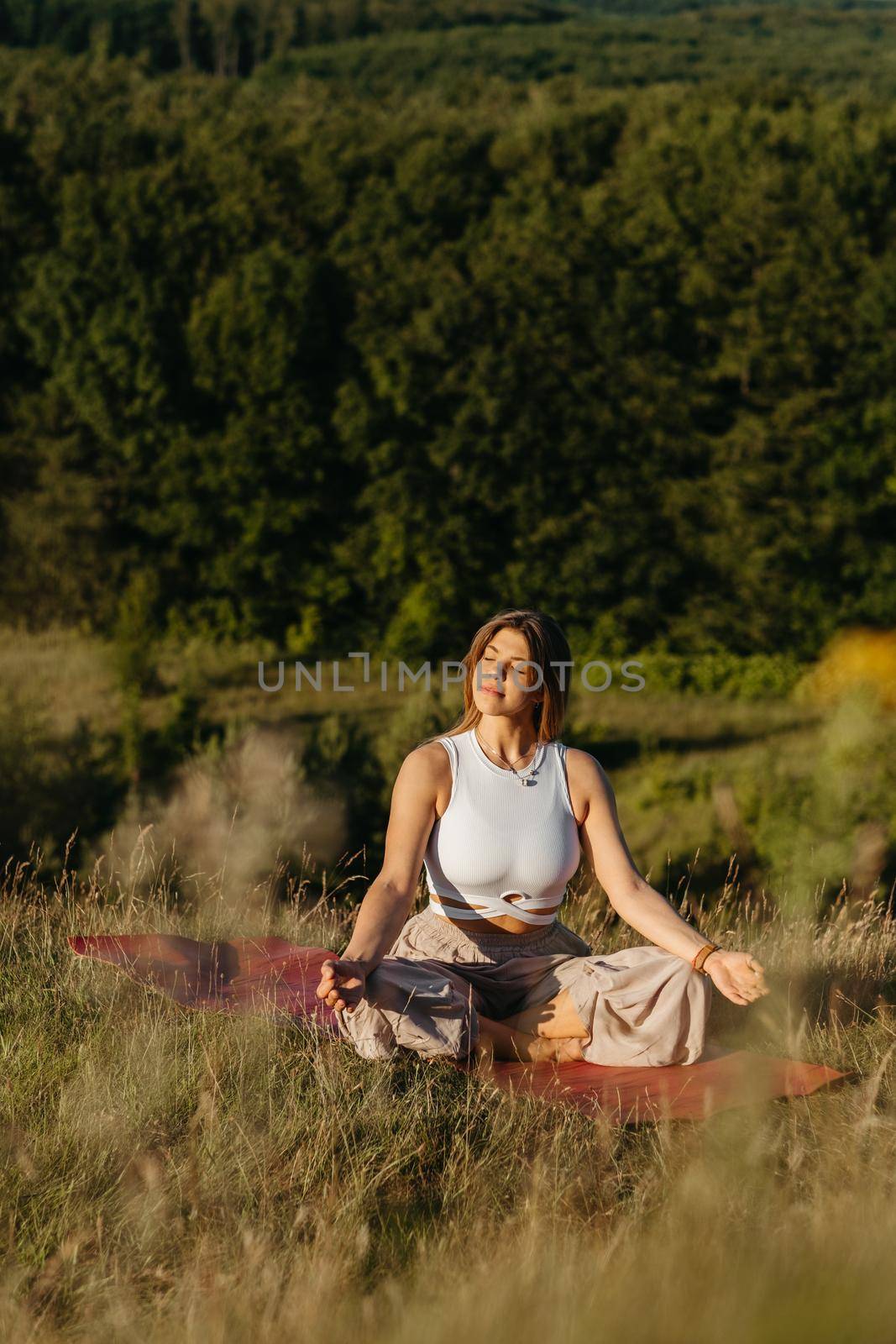 Young Woman Meditating on the Mat Outdoors at Sunset with Beautiful Landscape on Background