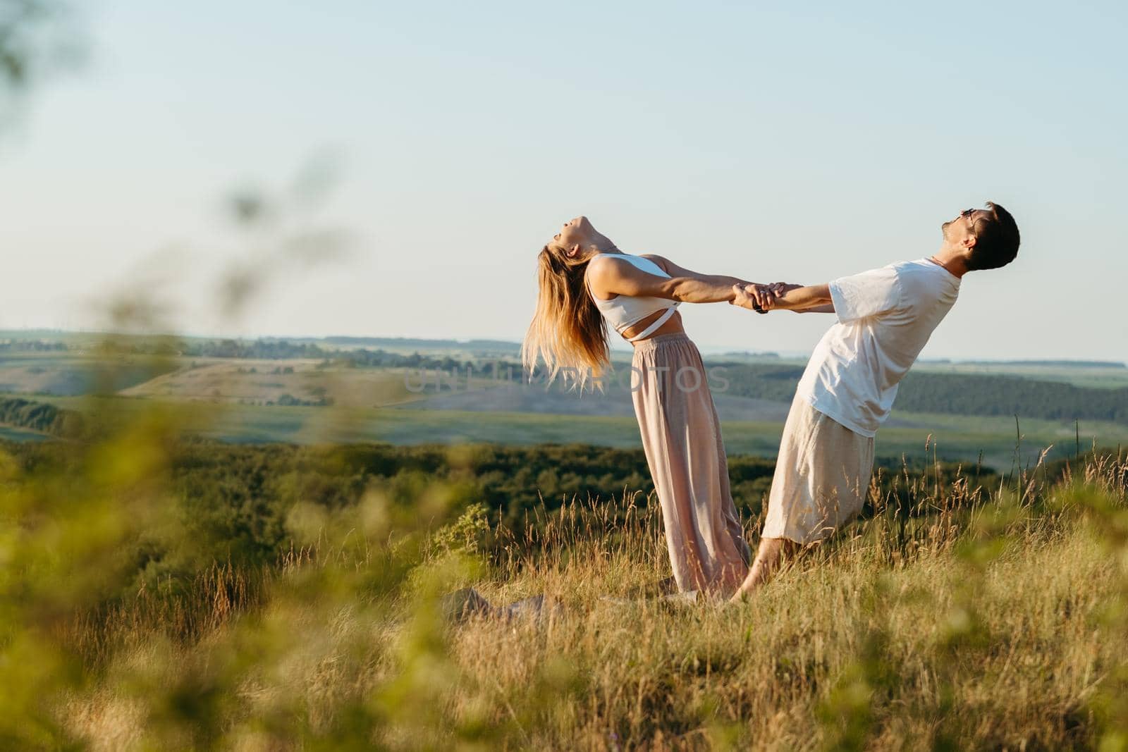 Young Adult Couple Holding by Hands, Man and Woman Practicing Yoga Outdoors with Scenic Landscape on Background at Sunset
