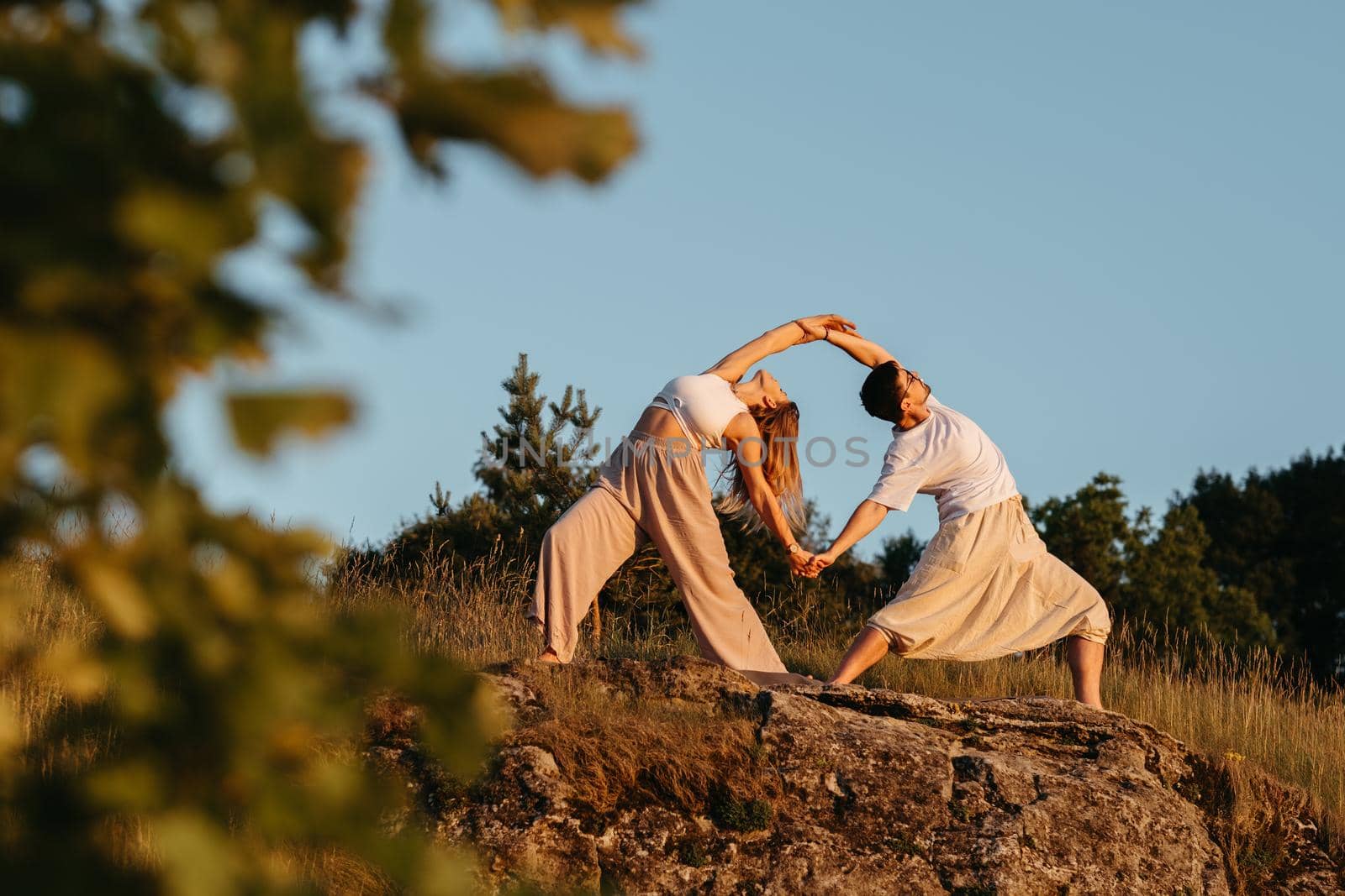Young Adult Couple Making Beautiful Pose, Man and Woman Practicing Yoga Outdoors on the Rocky Hill at Sunset by Romvy