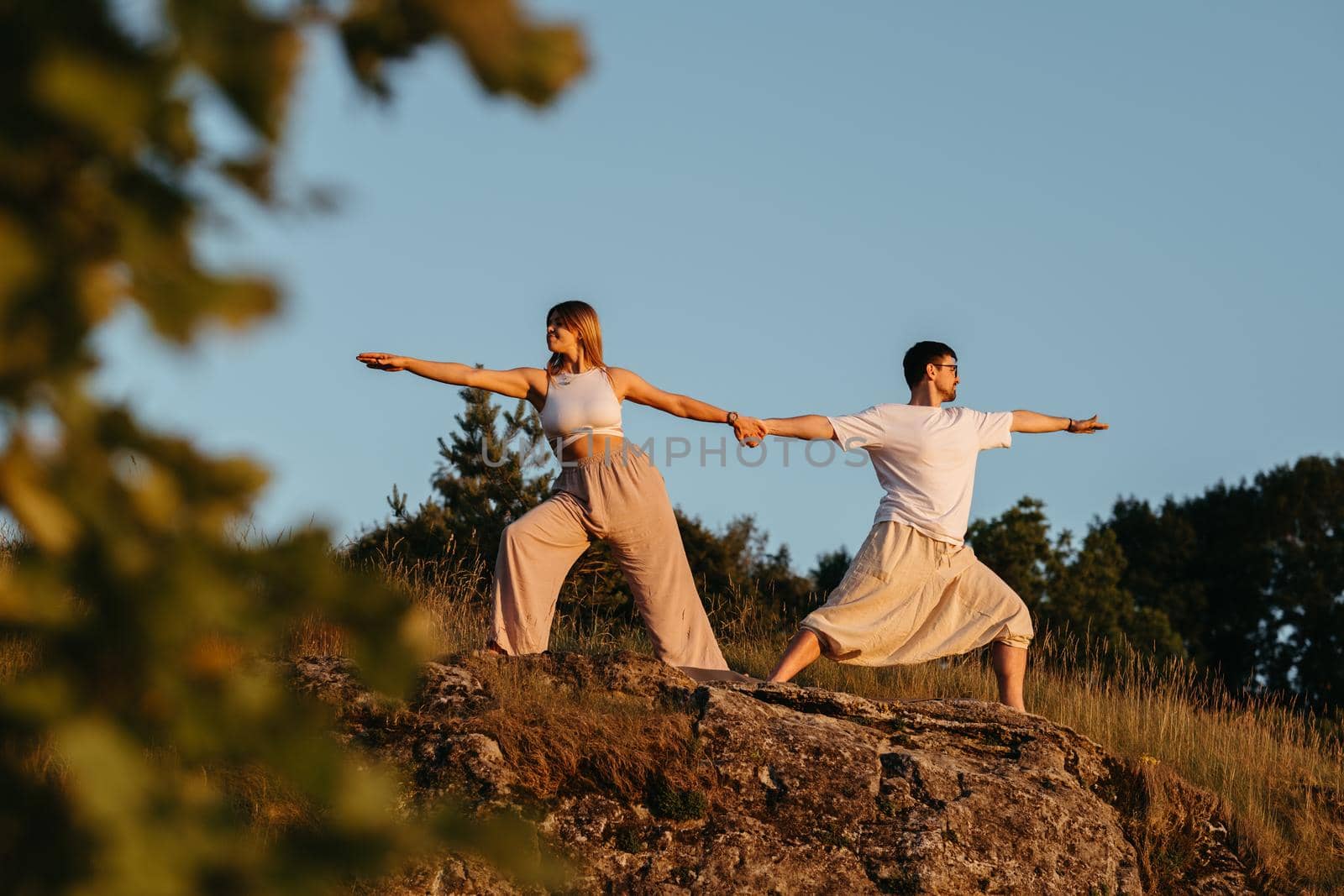 Young Adult Couple Holding by Hands, Man and Woman Practicing Yoga Outdoors on Rock at Sunset