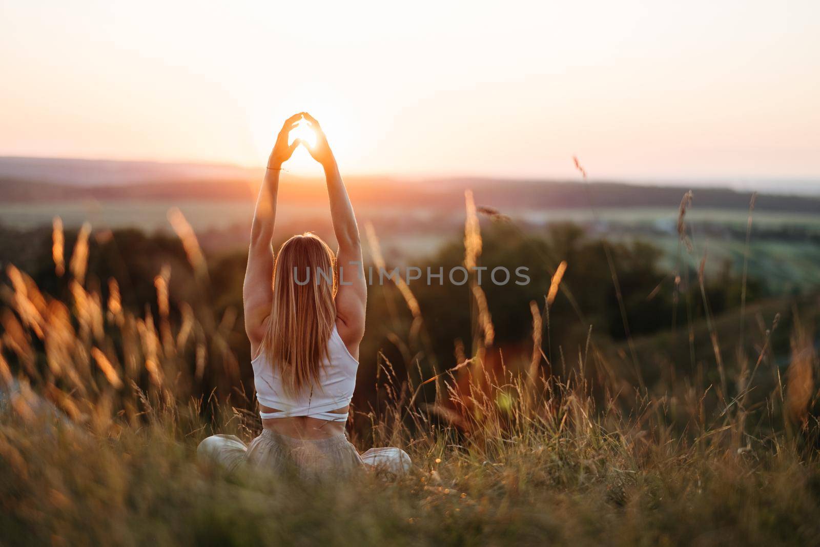 Back View on Woman Sitting in Meditation Yoga Pose and Catching Sun by Her Hands at Sunset Outdoors